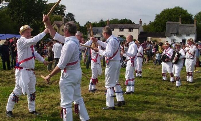 Morris dancers in white costumes perform with sticks at a village gathering